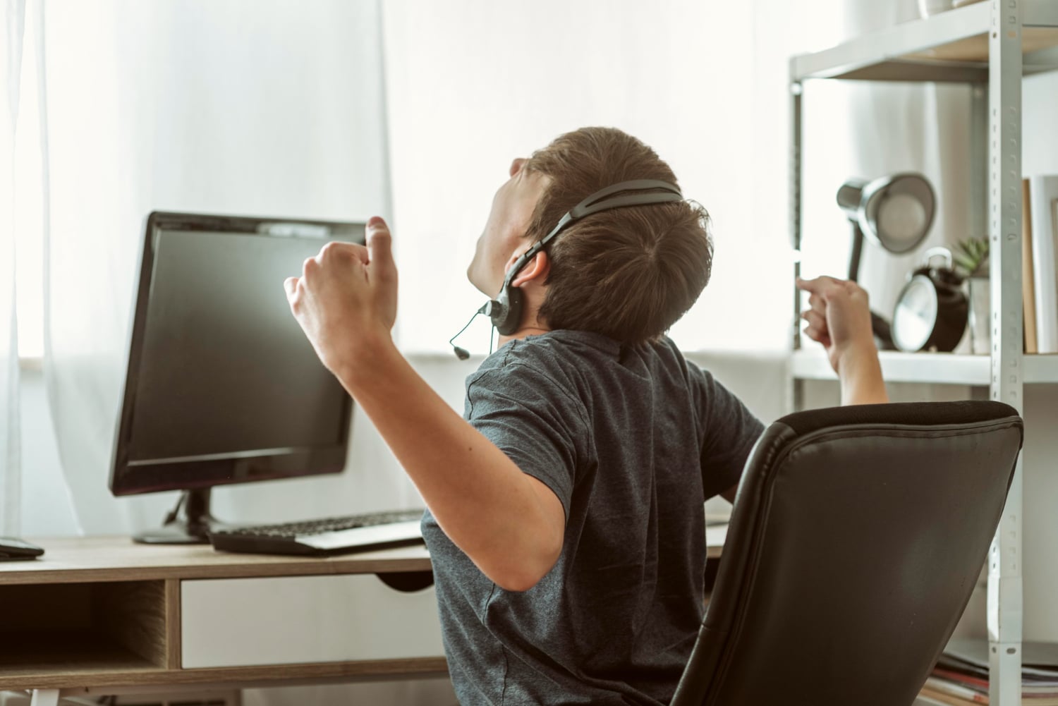 A man sitting in a chair with headphones on, speaking into a microphone while working at a desk with a computer monitor and other devices in an indoor office setting.