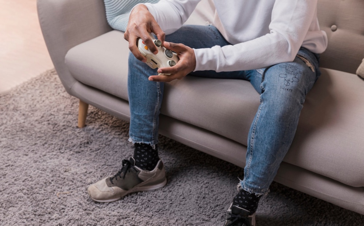 A person sits on a sofa, focused on a video game controller, wearing casual attire with ripped jeans and sneakers.