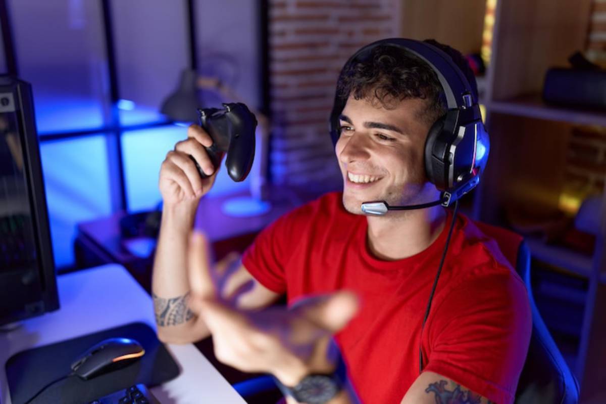 A gamer in a red shirt holds a game controller while wearing headphones, sitting in a dimly lit room with a computer setup.