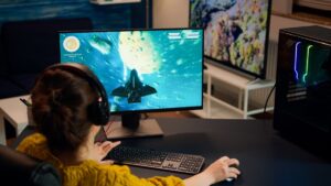 A woman gaming on a desktop computer, wearing headphones, with a spaceship displayed on the screen against a colorful space background.