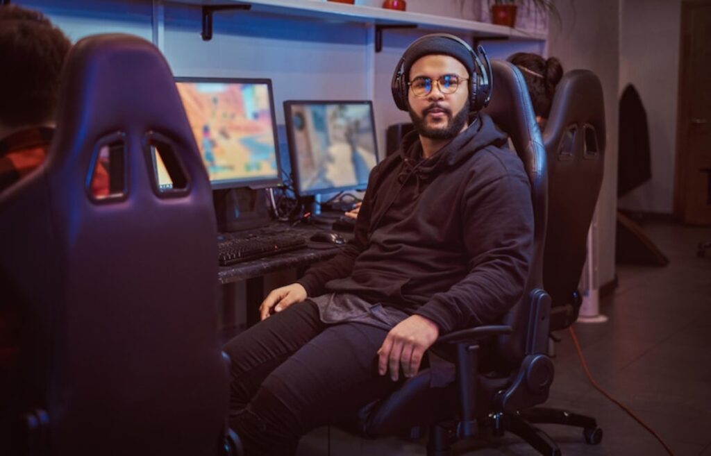 A person in a black hoodie and headphones sits relaxed in a gaming chair, surrounded by computer screens in a dimly lit room.