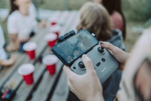 A person holds a drone controller with a smartphone attached, surrounded by red cups and friends enjoying a gathering outdoors.