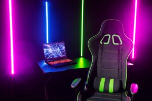 A gaming chair with green accents is placed by a desk with a laptop, surrounded by colorful neon light strips.