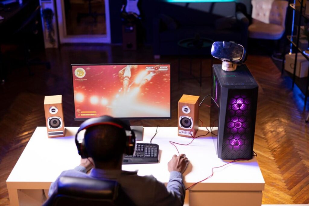 A gamer with headphones sits at a desk, focused on a colorful game displayed on the monitor, flanked by wooden speakers and a gaming PC.