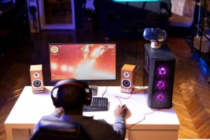 A gamer with headphones sits at a desk, focused on a colorful game displayed on the monitor, flanked by wooden speakers and a gaming PC.