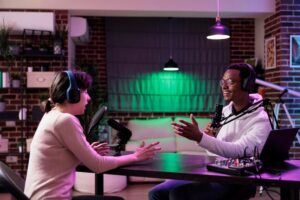 A podcast recording session in a cozy studio, featuring two people animatedly discussing at a table with microphones and audio equipment.