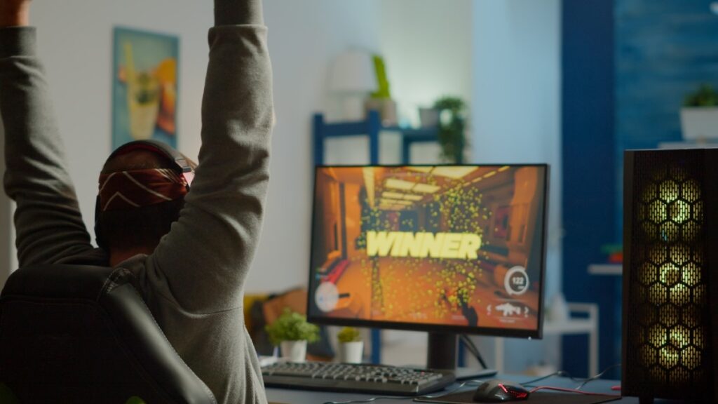 A gamer celebrates victory, raising arms in excitement, as a "WINNER" message displays on the monitor in a vibrant gaming setup.