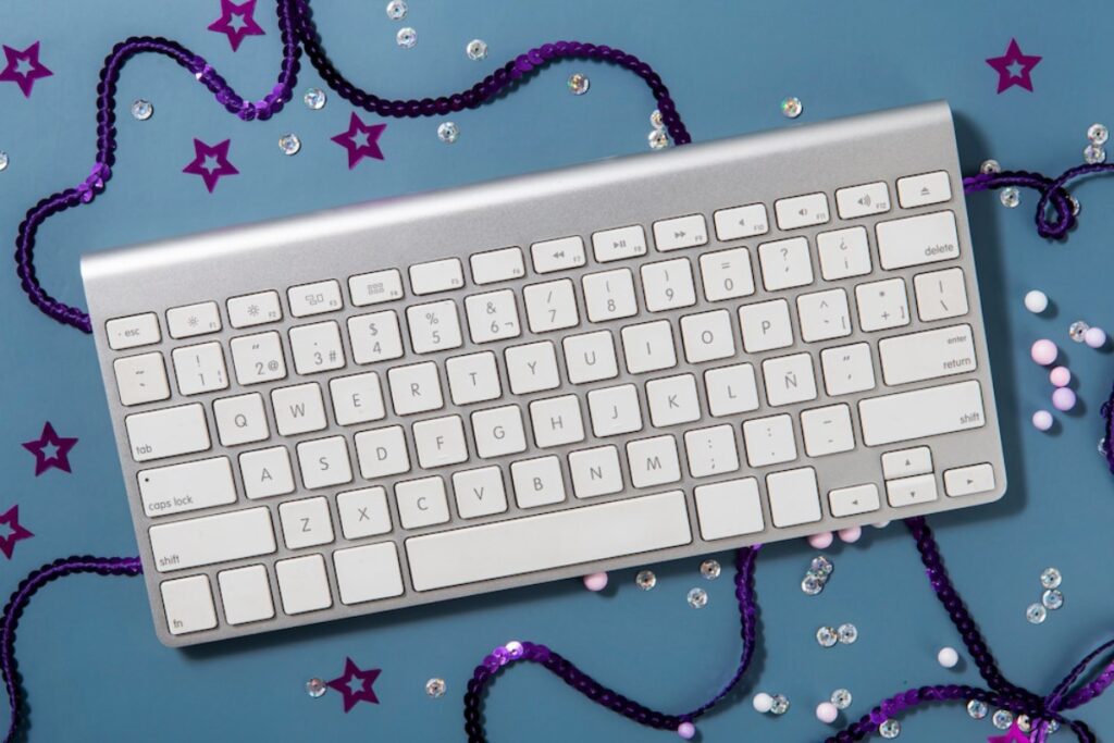An overhead view of a silver keyboard surrounded by colorful beads, sequins, and stars on a blue background.