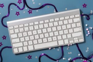 An overhead view of a silver keyboard surrounded by colorful beads, sequins, and stars on a blue background.