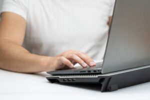 A person in a white shirt uses a laptop on a cooling stand, focused on the keyboard, with a clean white background.