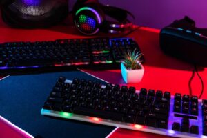 A colorful gaming setup featuring two backlit keyboards, headphones, and a small potted plant, all against a vibrant backdrop.