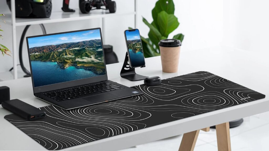 A sleek workspace featuring a laptop with a nature scene, a smartphone on a stand, a coffee cup, and a stylish desk mat with contour lines.