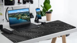 A sleek workspace featuring a laptop with a nature scene, a smartphone on a stand, a coffee cup, and a stylish desk mat with contour lines.