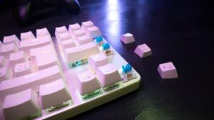 Close-up of a mechanical keyboard with white keycaps and blue switches, featuring some disassembled keys against a dark background.