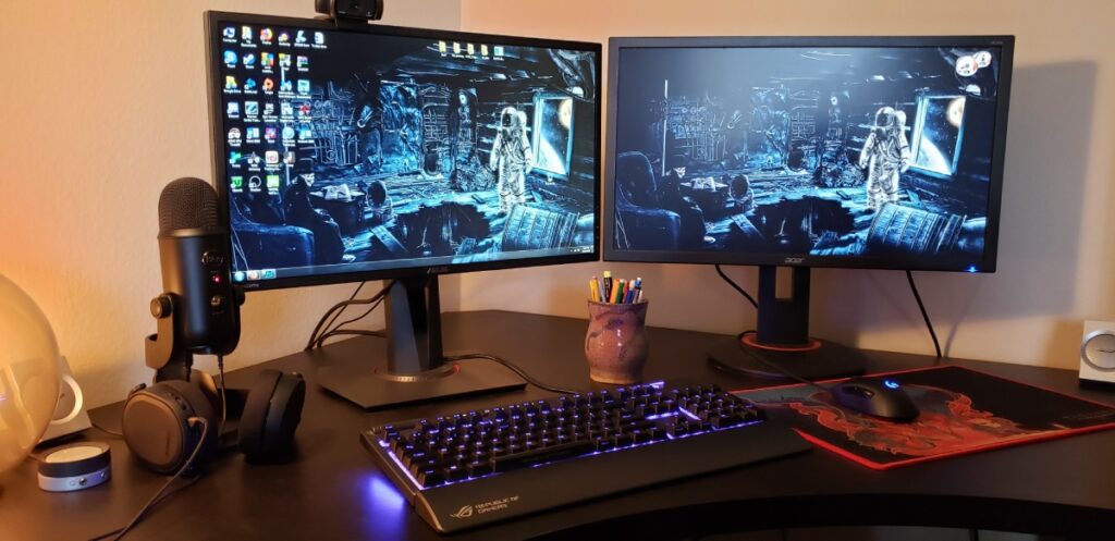 A dual-monitor setup featuring a gaming keyboard, headset, and microphone, with space-themed wallpapers and colorful desk accessories.
