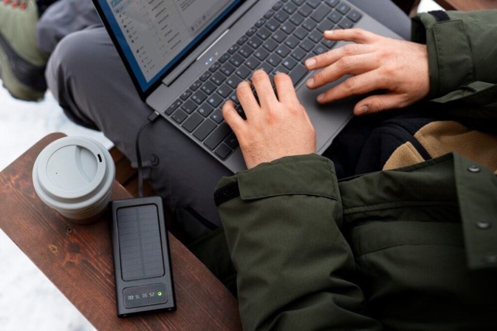 A person in a green jacket types on a laptop, with a coffee cup and solar charger nearby on a wooden surface.