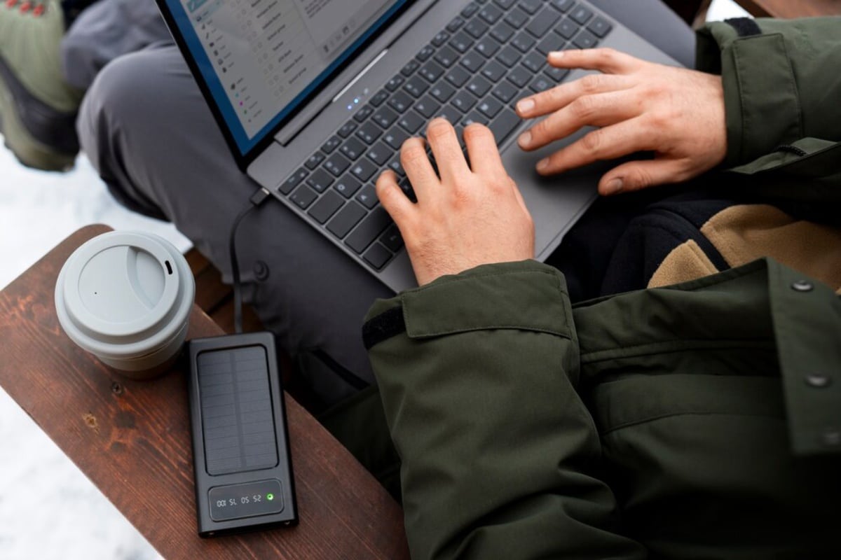 A person in a green jacket types on a laptop, with a coffee cup and solar charger nearby on a wooden surface.