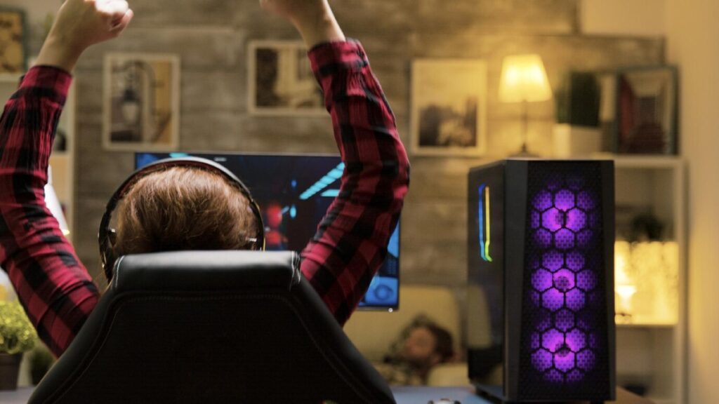 A gamer celebrates a victory with raised arms while sitting at a computer desk, illuminated by a colorful, lit PC tower.
