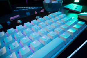 A close-up of a white keyboard illuminated with blue and pink lights, highlighting the keys and their labels on a dark desk.