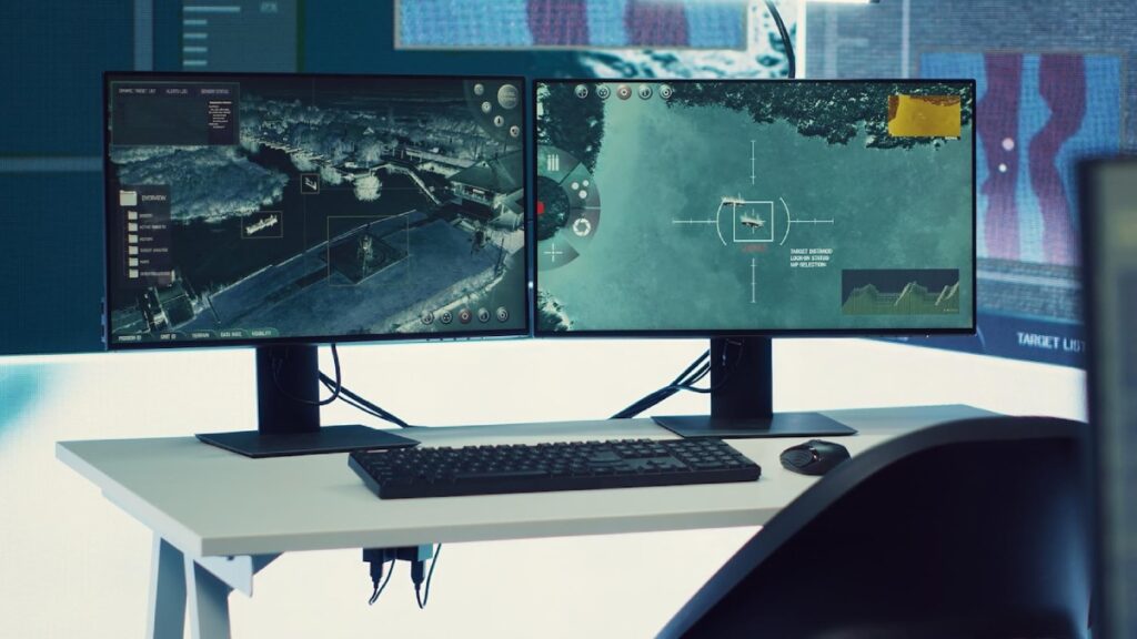 A dual-monitor setup displays tactical surveillance and targeting software, with a keyboard and mouse on a modern workspace.