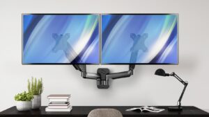 Dual monitors mounted on a sleek, adjustable arm above a dark desk with books, plants, and a desk lamp in a modern workspace.
