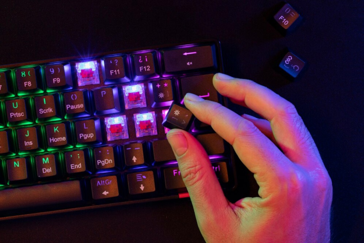 A hand is about to remove a keycap from a backlit keyboard with colorful LED lights under the transparent switches.