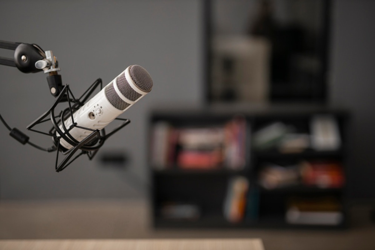Close-up of a white microphone mounted on a black arm, with a blurred bookshelf in the background. Ideal for podcasting or broadcasting.