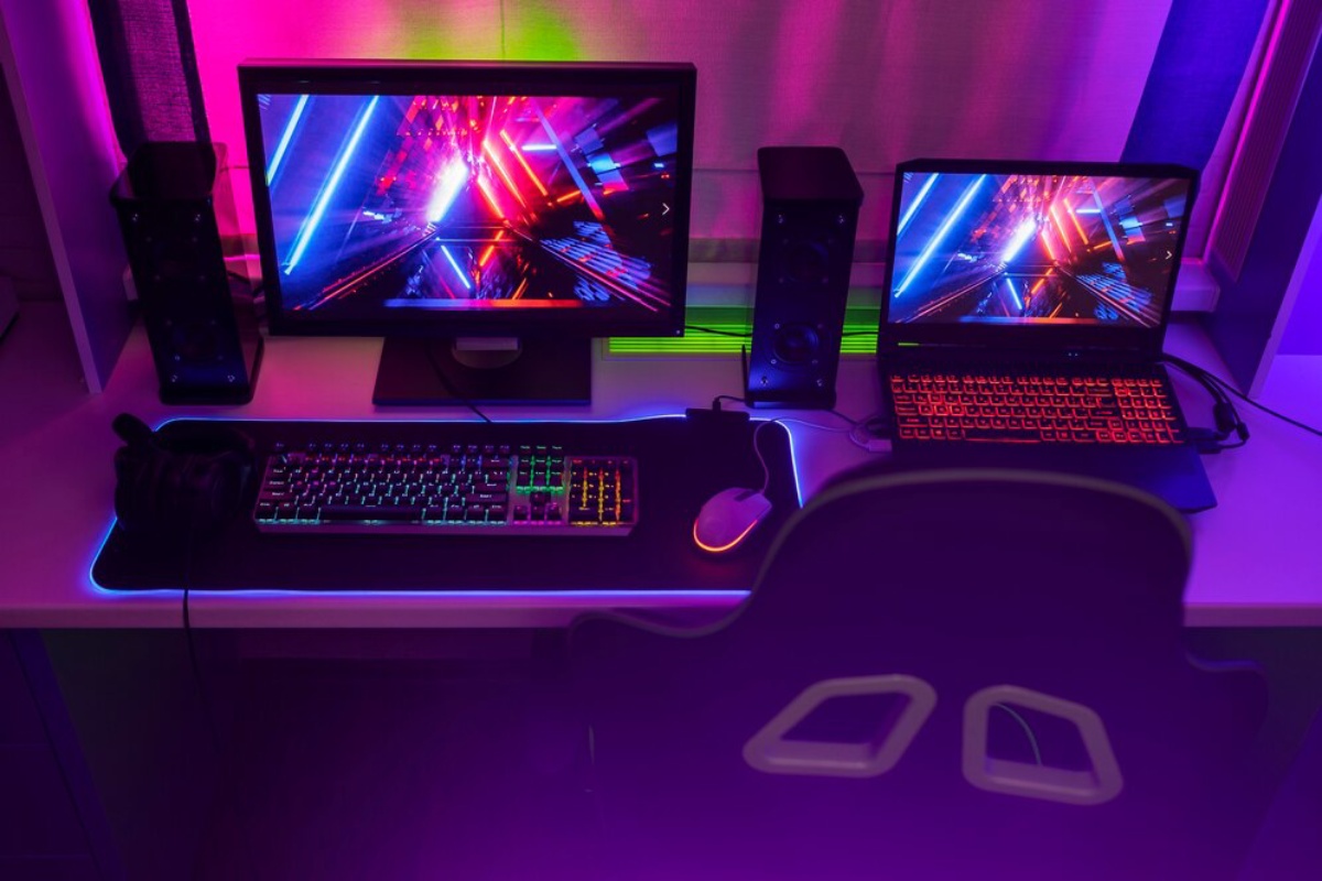 A vibrant gaming setup featuring two screens with colorful visuals, a sleek keyboard, mouse, headphones, and speakers in neon lighting.