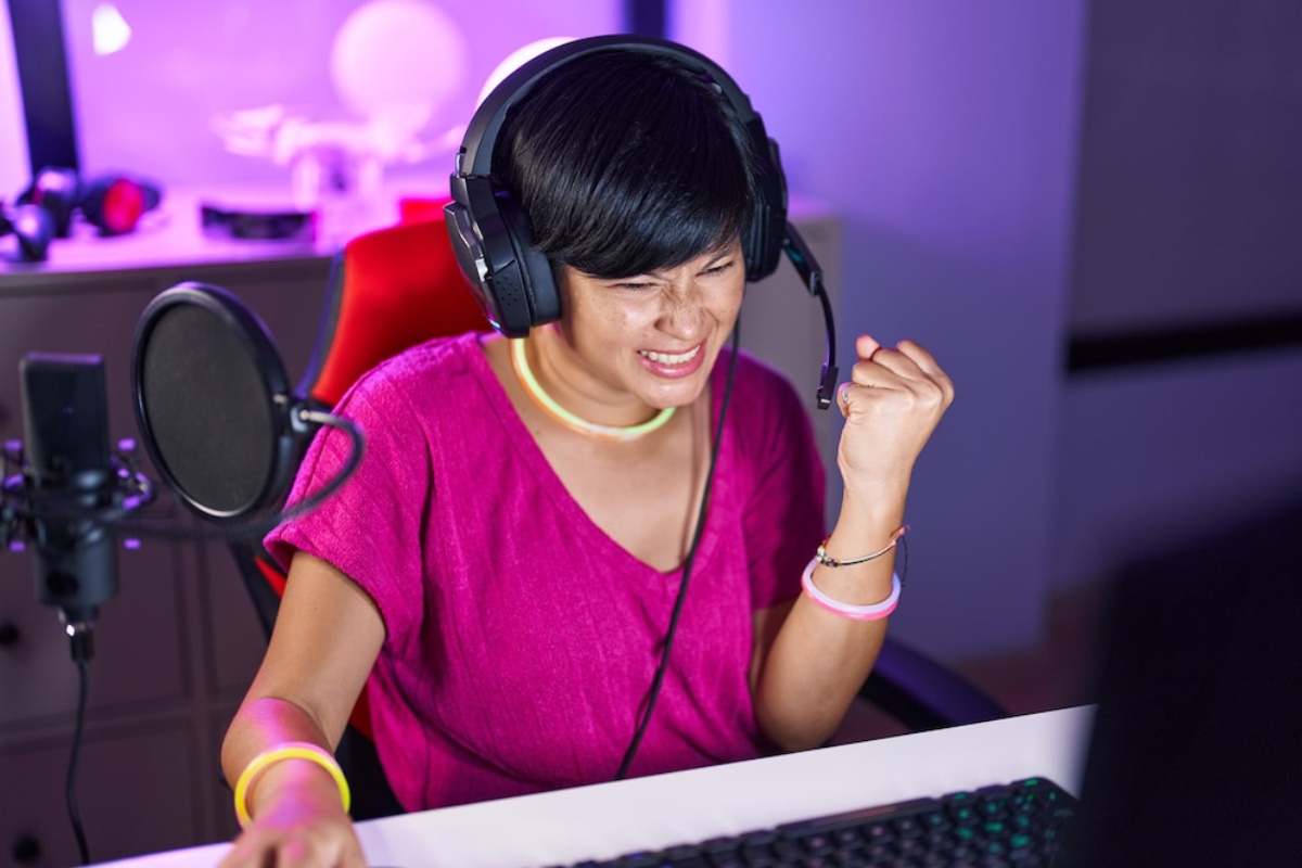 A person wearing headphones and a headset passionately gestures while gaming at a computer, surrounded by colorful ambient lighting.