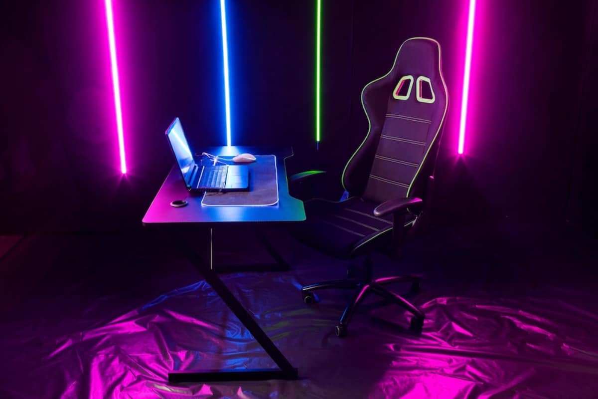 A sleek gaming setup featuring a modern desk, a laptop, and a racing-style gaming chair, illuminated by colorful neon lights.