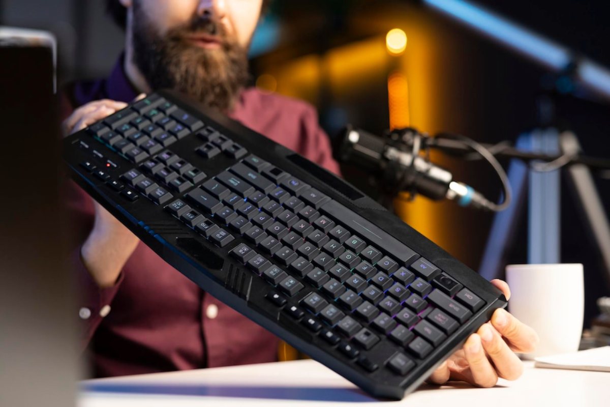 A person holds a backlit black keyboard, with a white mug and microphone visible on a well-lit desk.