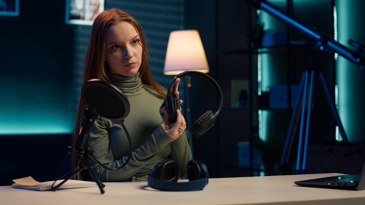 A person in a green sweater holds a headset in front of a microphone on a desk, surrounded by a modern, softly lit setup.