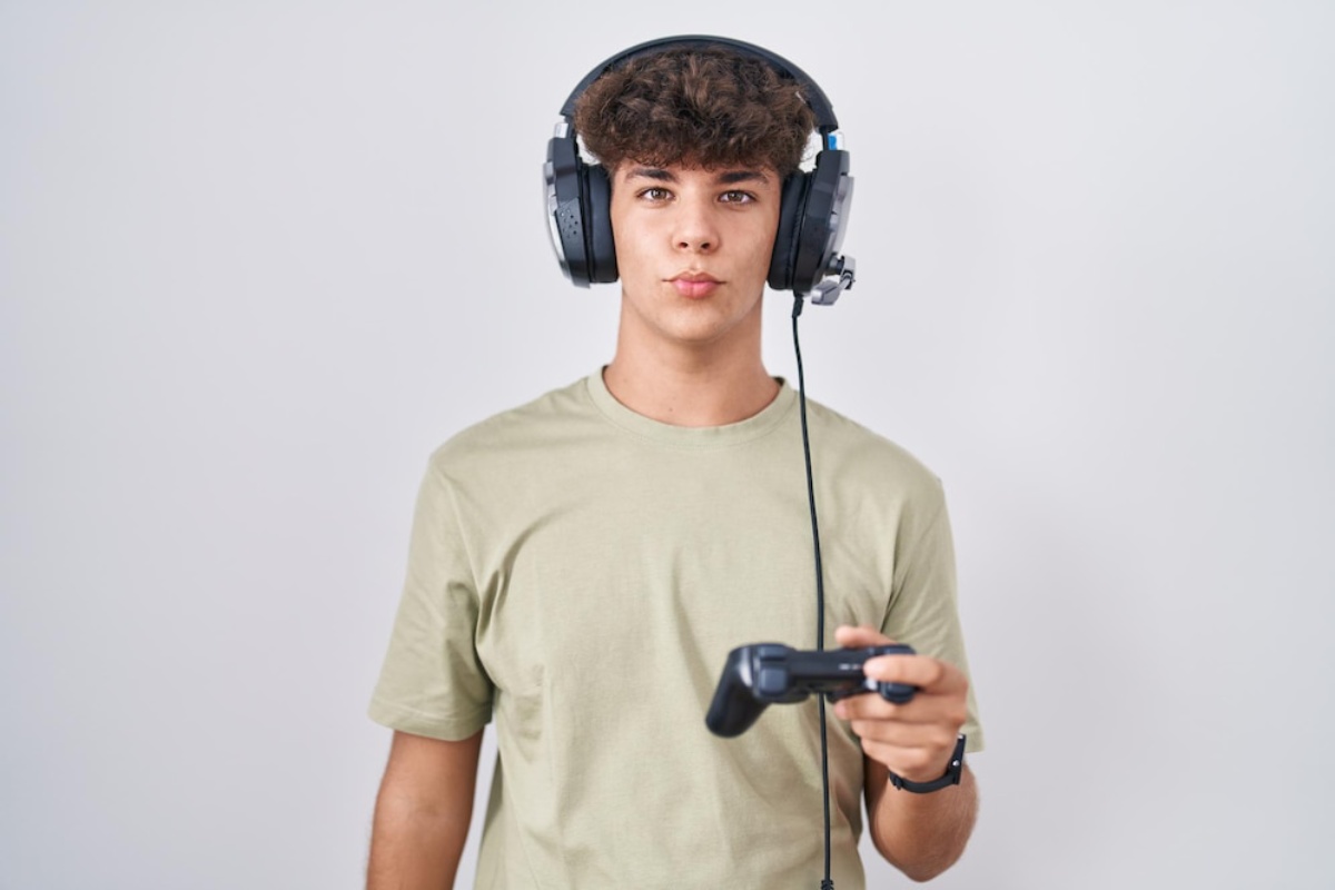 A person wearing large headphones holds a video game controller, dressed in a light green t-shirt, set against a plain background.