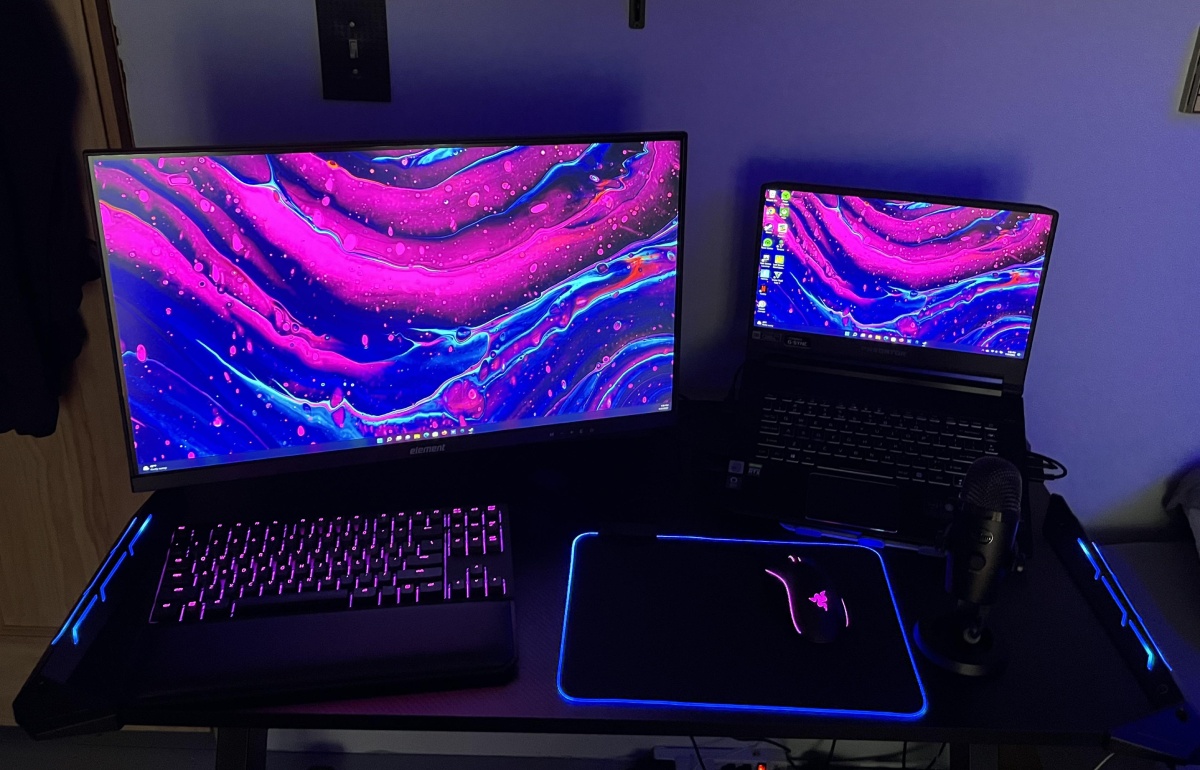 A gaming setup featuring a large monitor and a laptop, both displaying vibrant, colorful abstract wallpapers, with RGB backlit keyboard and mouse.