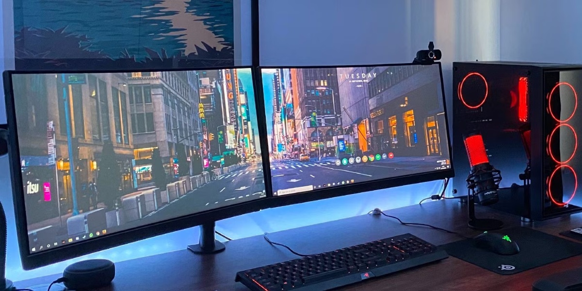 A dual-monitor setup displays a cityscape wallpaper, with a gaming PC, microphone, and RGB lighting on a wooden desk.