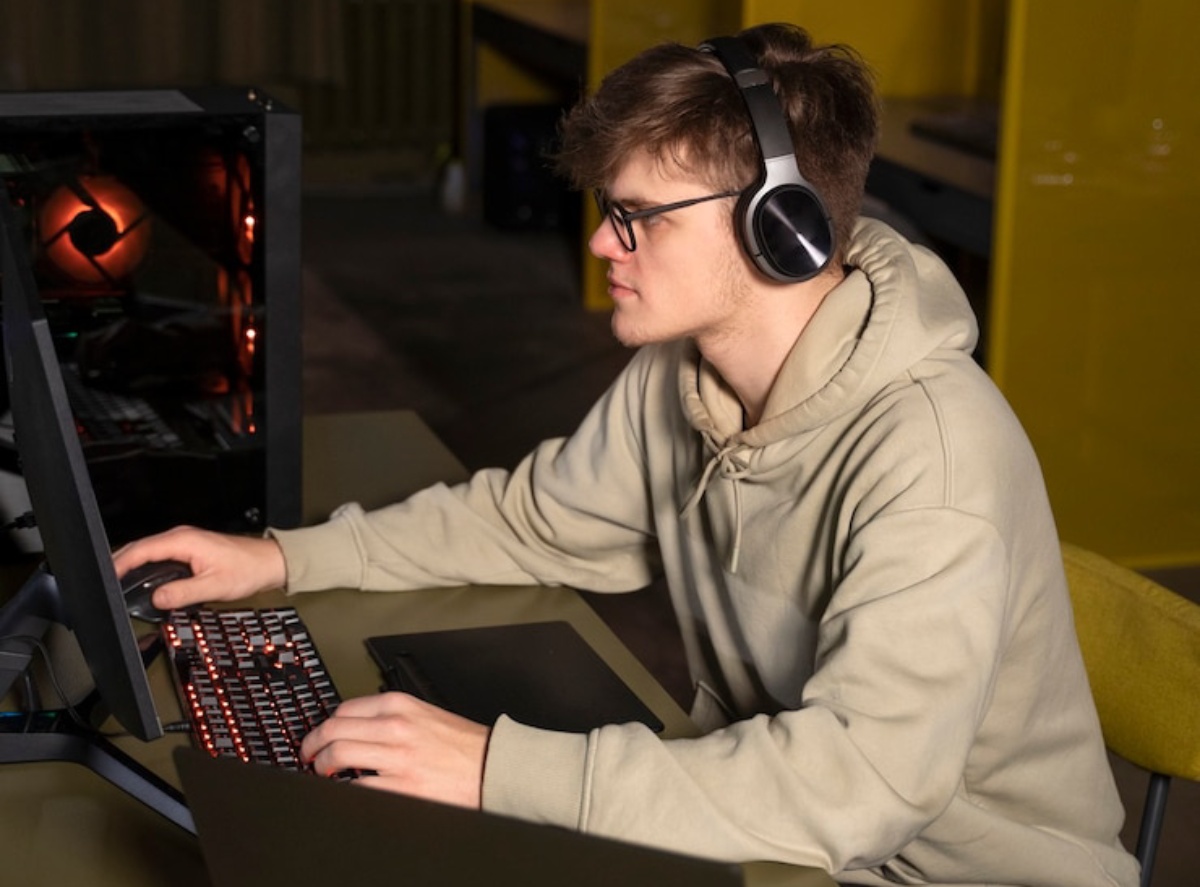 A person in a beige hoodie uses a gaming keyboard and mouse, wearing headphones, in a tech-filled environment with colorful lighting.