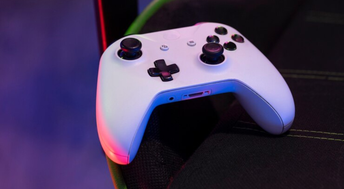 Close-up of a white gaming controller resting on a black seat, illuminated with colorful LED lights in a dimly lit environment.