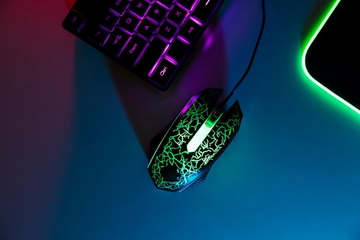 A colorful gaming mouse with a cracked design, illuminated in green and purple, sits beside a backlit keyboard on a gradient background.