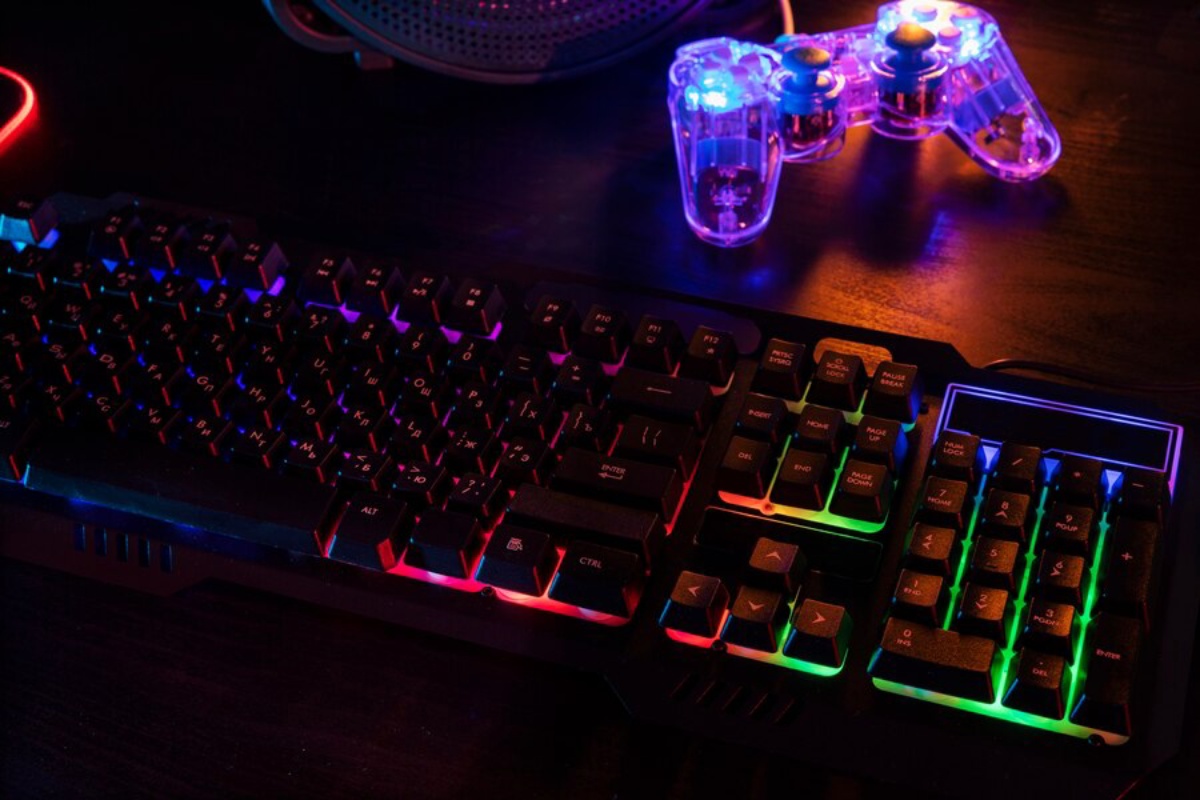 A backlit gaming keyboard with vibrant RGB lighting and a transparent game controller on a dark wooden surface.