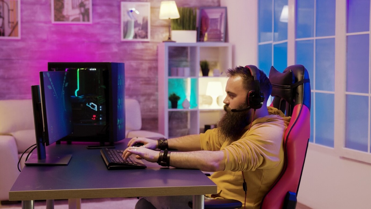 A person wearing headphones sits at a desk, gaming on a computer with colorful lighting in a cozy, modern room.