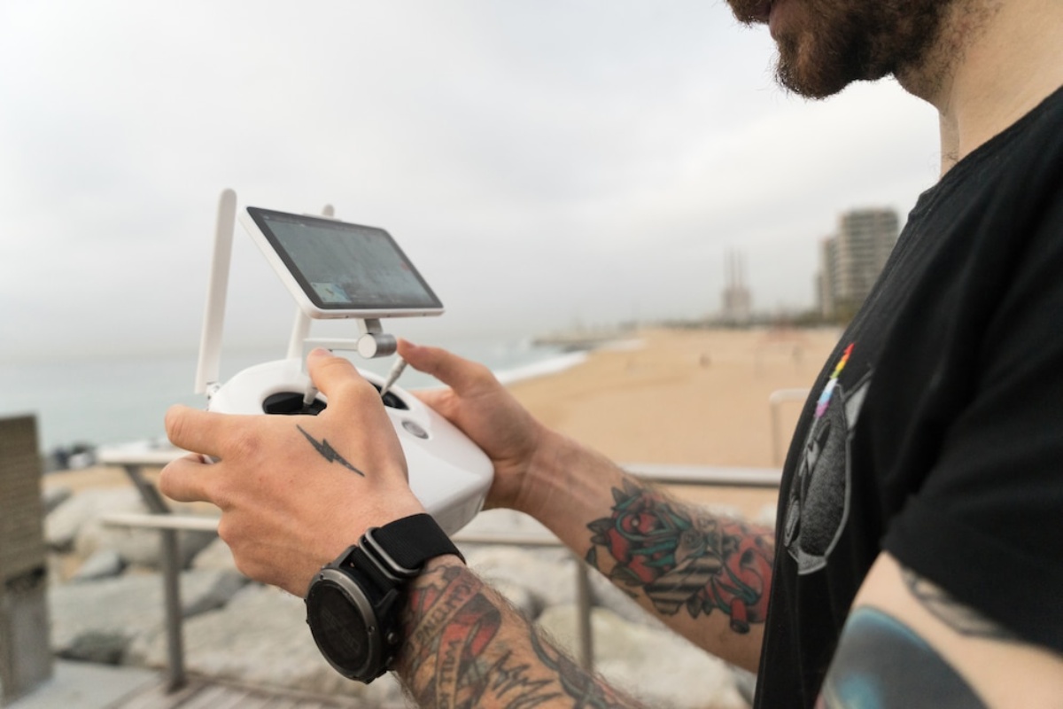 A person with tattoos controls a drone using a remote with a screen, overlooking a beach and cloudy sky.