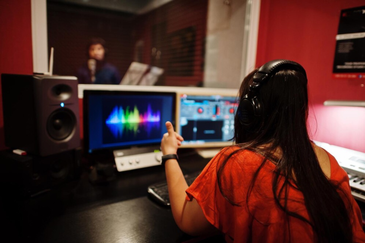 A woman in headphones controls audio equipment in a recording studio, focusing on colorful sound wave visuals on her computer screens.