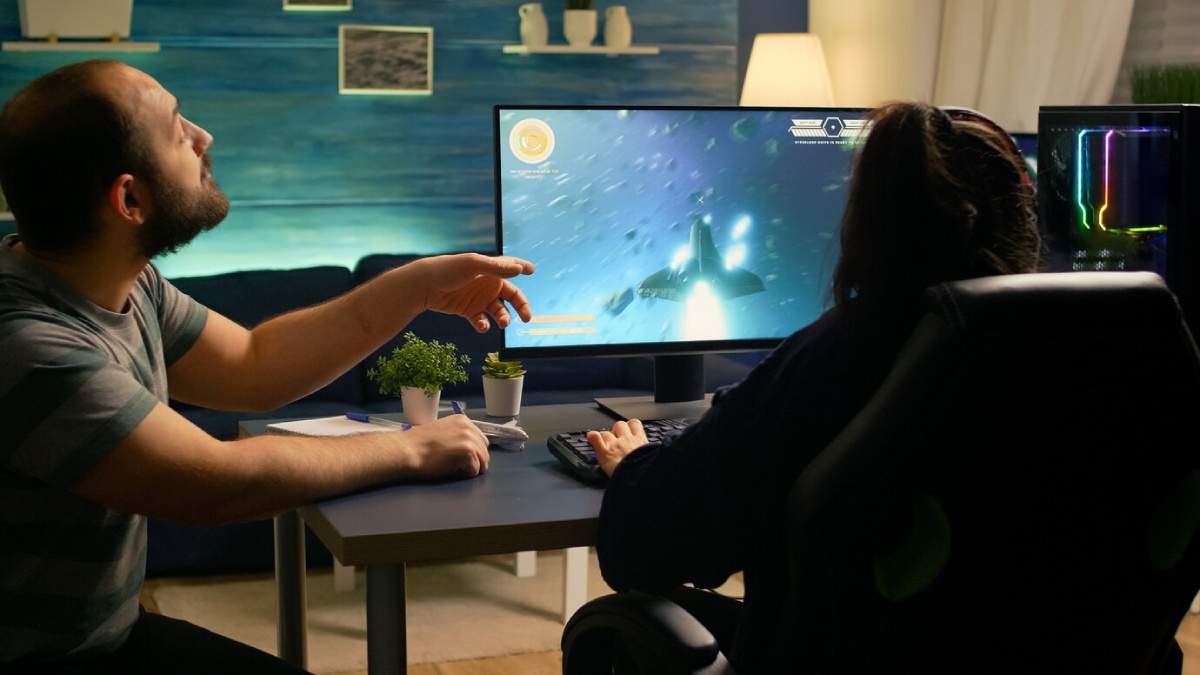 Two people are engaged in gaming at a desk, with one pointing at a screen showing a spaceship in a colorful space scene.