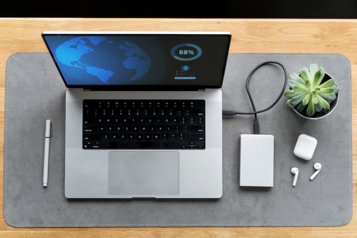 A laptop displaying a world map and battery percentage, next to a portable hard drive, headphones, and a green succulent on a gray mat.