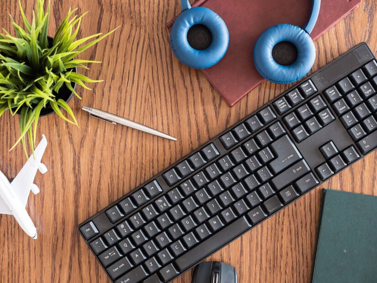 Top view of a desk with a black keyboard, mouse, headphones, pen, small plant, airplane model, and books on a wooden surface.