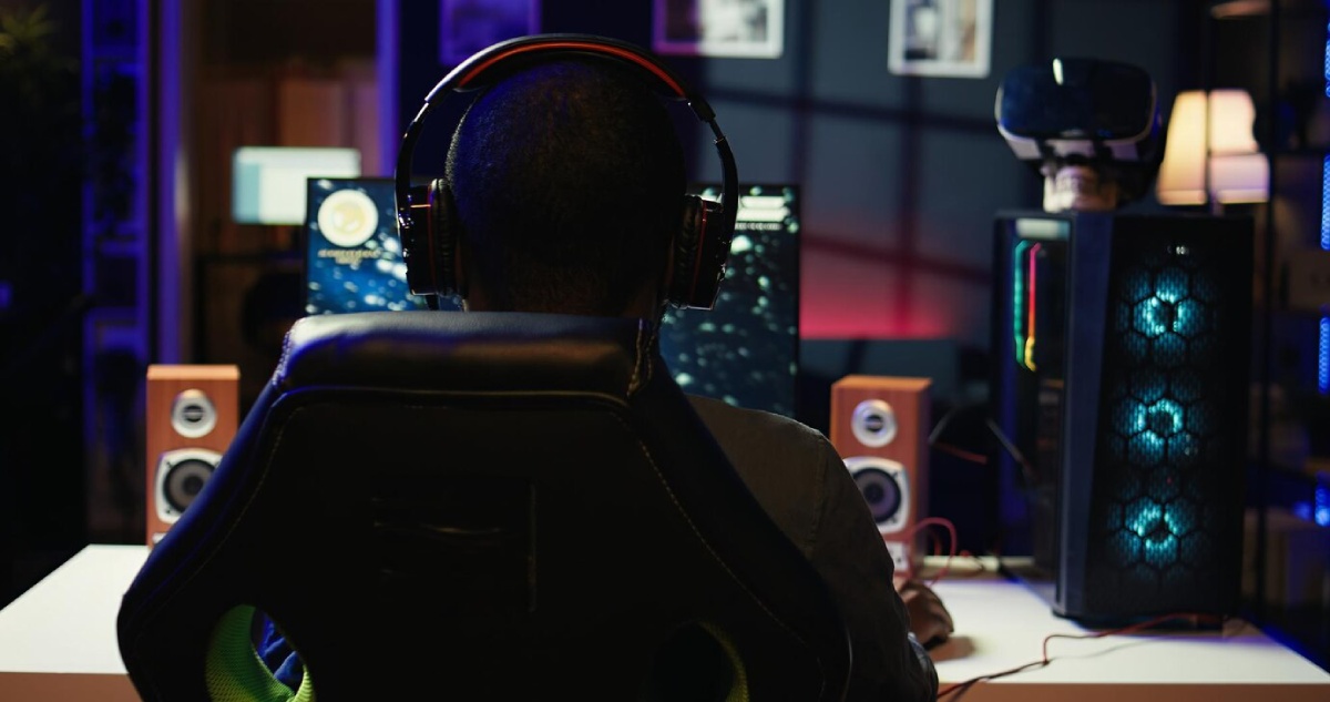 A gamer in headphones sits in a high-backed chair, focused on a monitor, with vibrant lighting and speakers in the background.