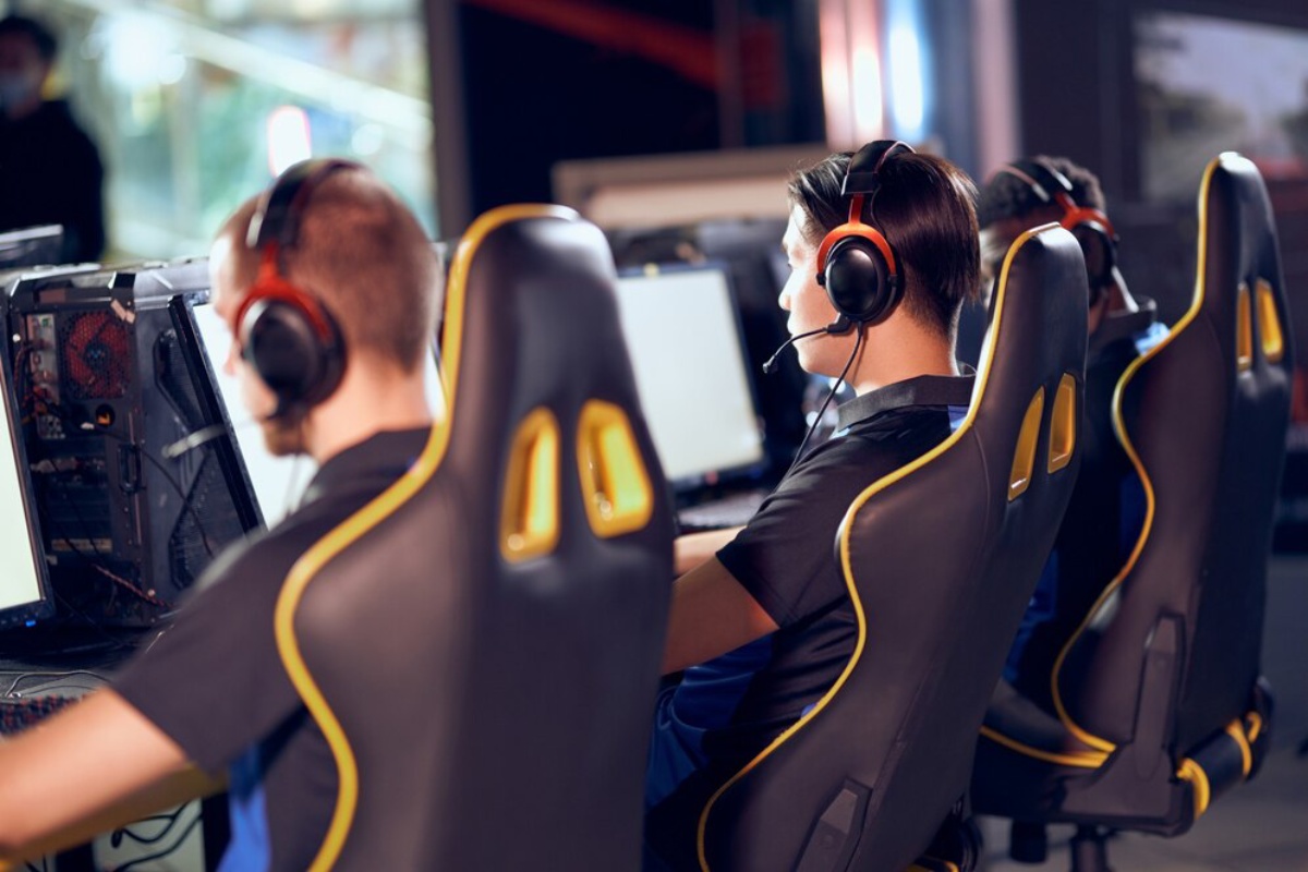 Four gamers, focused and wearing headsets, sit in ergonomic gaming chairs at a row of computers in a gaming center.