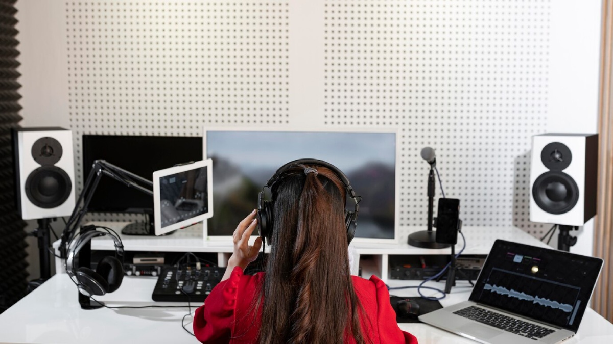 A woman in a red blouse listens through headphones in a modern audio studio, surrounded by monitors and recording equipment.