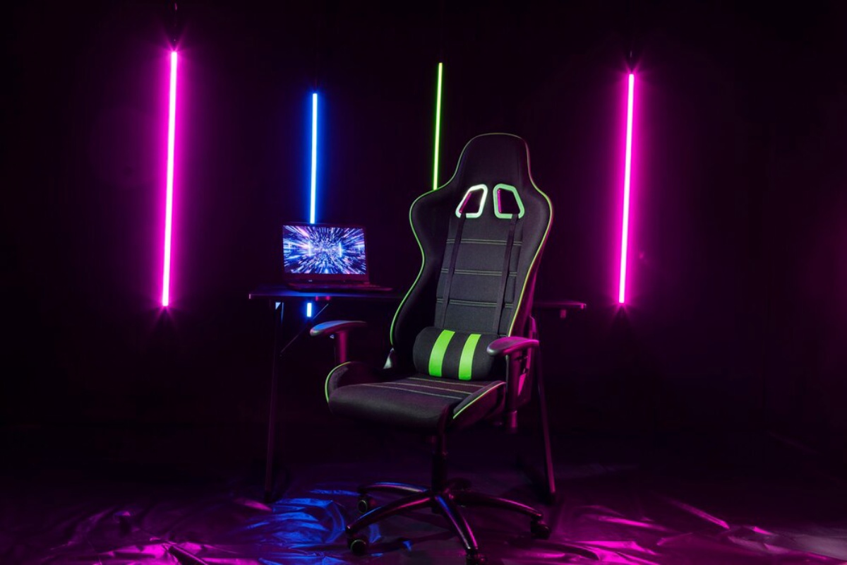 A gaming chair with green accents sits beside a laptop on a desk, illuminated by pink and blue neon lights in a dark setting.