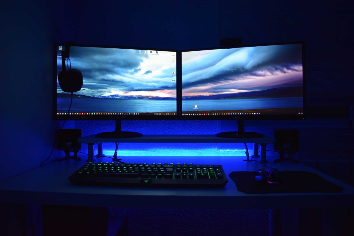 A dual-monitor setup with vibrant blue lighting, a green backlit keyboard, and a gaming mouse on a sleek desk in a dimly lit room.